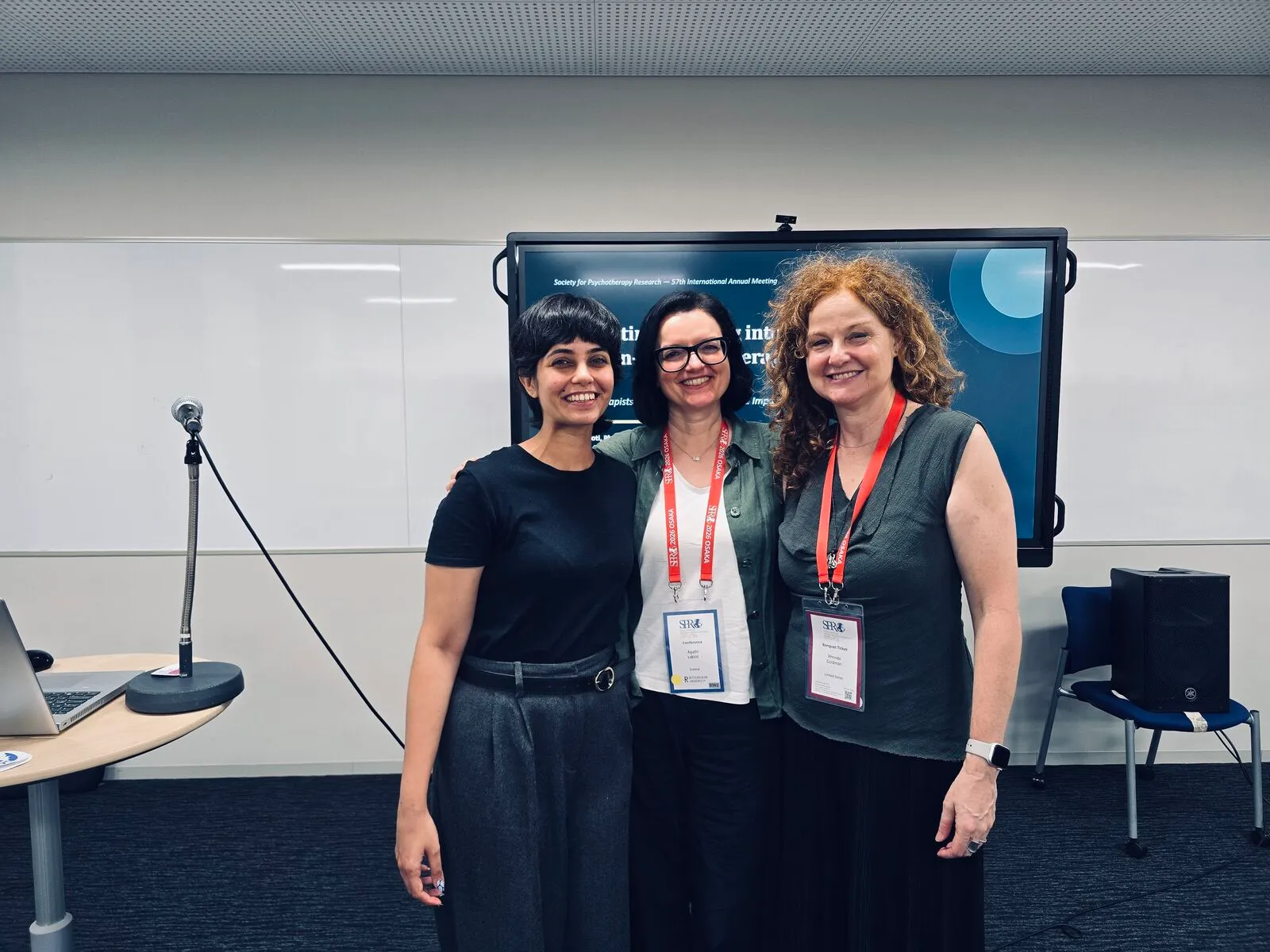 Three women pose together in front of a presentation screen at the conference